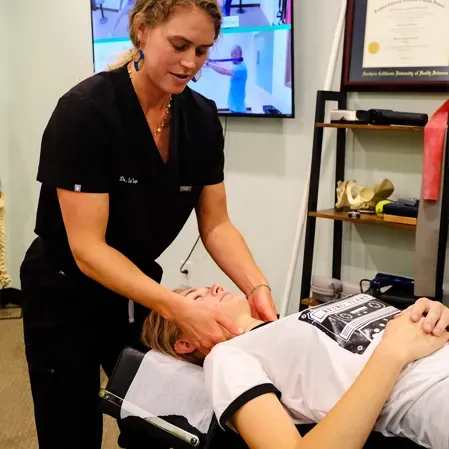 Person lying on a treatment table while another supports and adjusts the neck, with clinic equipment in background.