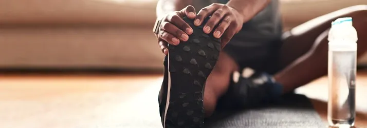 Person seated on a mat holding one foot with both hands, showing sole of shoe, with a water bottle beside.
