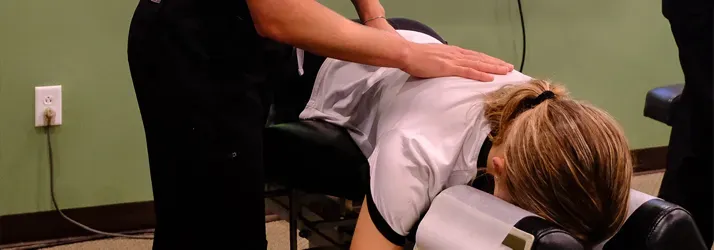 Person lying face down on a treatment table while another presses one hand on the upper back near the shoulder area.