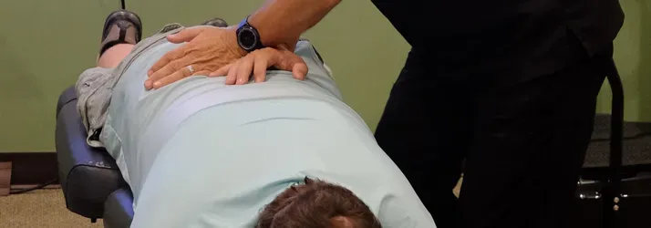 Person lying face down on a treatment table while another applies pressure with both hands to the lower back.
