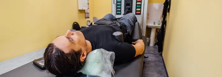 Person lying on a treatment table with head supported and a strap across legs in a clinical room with equipment.