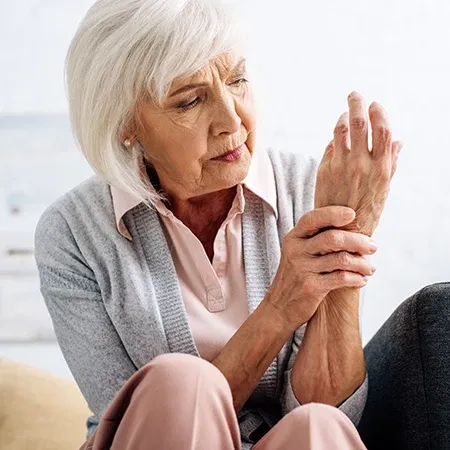 Person seated indoors holding and pressing one wrist with the other hand, wearing light clothing against a neutral background.