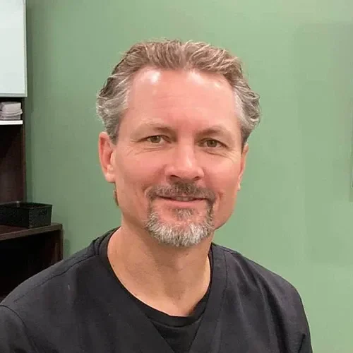 Dr. Greg Grochowski is seated in a clinical room wearing dark scrubs, with cabinets, stool, and equipment visible against a green wall.